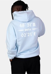 Ljusblå hoodie med en stor vit grafik på ryggen som visar koordinater och ordet "paris." Mjukt material, avslappnad passform, justerbar huva.