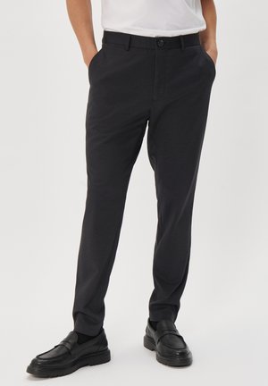 Pantalones negros de corte slim con una textura suave, que cuentan con bolsillos laterales y un cierre de botón. Se llevan con zapatos negros tipo mocasín y una camisa blanca.