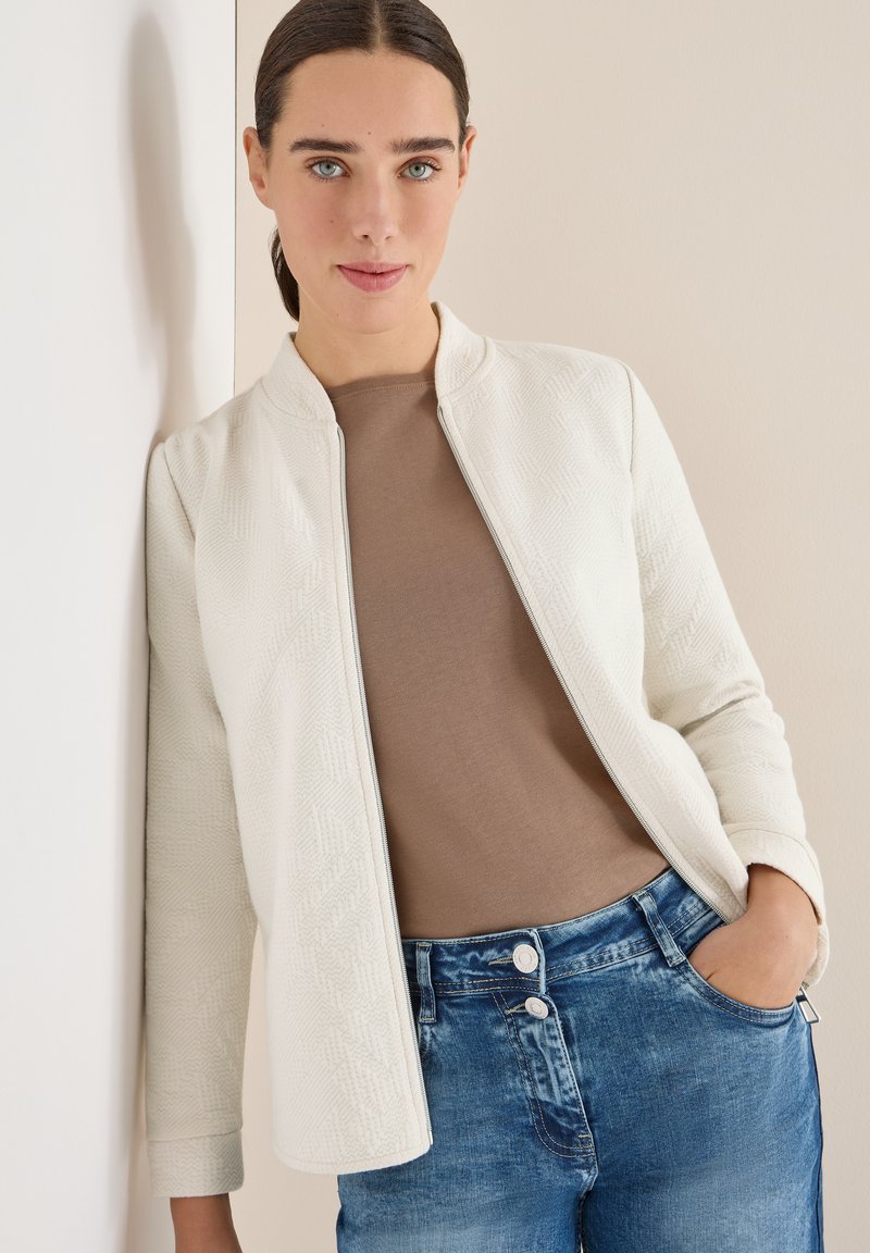 Bílá texturovaná bunda s mandžestráckým límcem, doplněná hnědým topem a modrými džínami. Bunda má geometrický vzor a zapínání na zip.