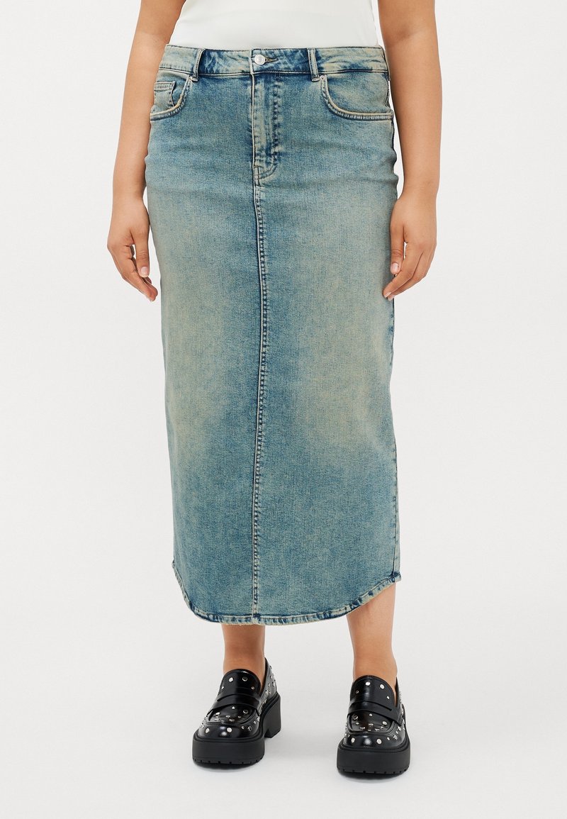 Femme portant une jupe midi en denim bleu et des mocassins à plateforme noirs cloutés, se tenant sur un fond blanc.