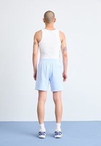 Man står på blått golv mot vit vägg, iklädd vit linne, ljusblå shorts, vita strumpor och vita sneakers, och visar bakifrån.