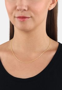 Kvinna som bär ett delikat vridet guldhalsband över en svart rundhalsad topp, som visar detaljer av hals och nedre ansikte.