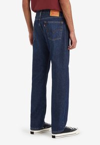 Mörkblå denimjeans med rak skärning, med en läderlapp på bakre midjan och dubbla bakfickor. Svarta sneakers synliga nedanför.