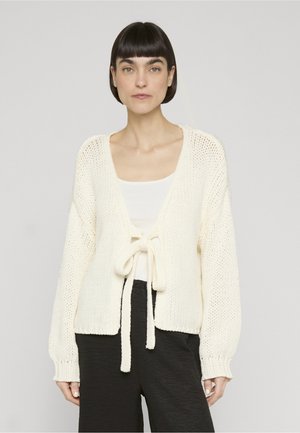 Kvinde med kort mørkt hår iført en off-white strikket cardigan bundet foran over en hvid top og sorte bukser, stående mod en ensfarvet baggrund.