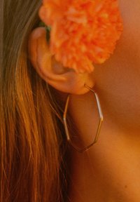 Metallisches sechseckiges Creolen-Ohrring mit glänzender Goldoberfläche, im Ohr getragen; eine orangefarbene Blume verdeckt teilweise die Sicht. Braunes Haar sichtbar.