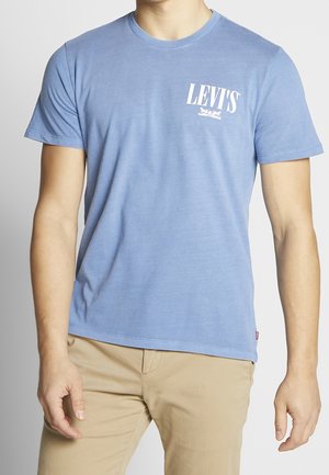 Mężczyzna ubrany w jasnoniebieski T-shirt Levi's z krótkim rękawem i białym logo na piersi oraz beżowe spodnie, na jednolitym tle.