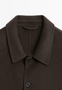 Cappotto in lana marrone con colletto, chiusura a bottoni e una morbida trama in pile. Il tessuto appare spesso e caldo, ideale per il clima più fresco.