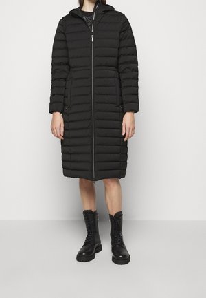 Femme portant un manteau matelassé noir longueur genou avec une fermeture éclair à l'avant et une capuche, associé à des bottines noires à lacets.