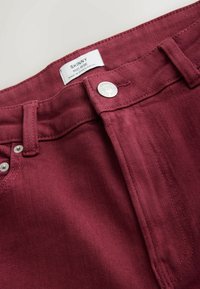 Burgundy skinny jeans featuring mid-rise waist, silver button, and contrasting stitching. Smooth texture with a slight sheen.