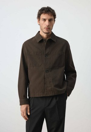 Hombre con pelo corto y rizado que lleva una chaqueta marrón oscura abotonada con bolsillos delanteros y pantalones negros, de pie contra un fondo liso y claro.