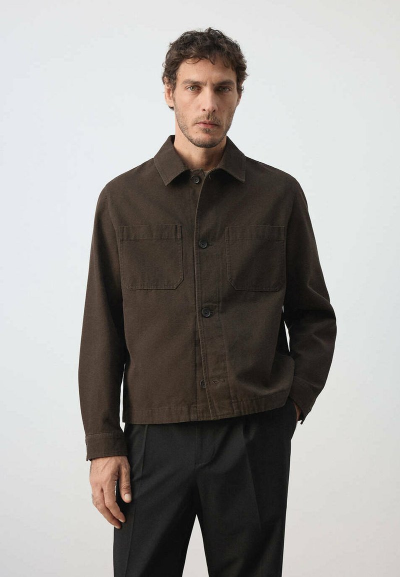 Homme aux cheveux courts et bouclés portant une veste boutonnée marron foncé avec des poches avant et un pantalon noir, debout devant un fond clair uni.