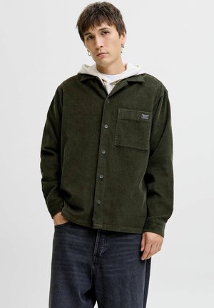 Camisa de pana en verde oscuro con cuello, bolsillo frontal y cierre de botones. Llevada sobre una sudadera blanca, combinada con jeans oscuros.