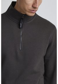 Pullover grigio scuro con colletto alto e mezza zip. Texture liscia, design aderente, dotato di un sottile etichetta con logo sulla zip.