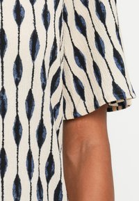 Vêtement à manches courtes en tissu texturé présentant une base crème avec des motifs en forme de goutte d'eau bleus et noirs, et une bordure rayée contrastante.