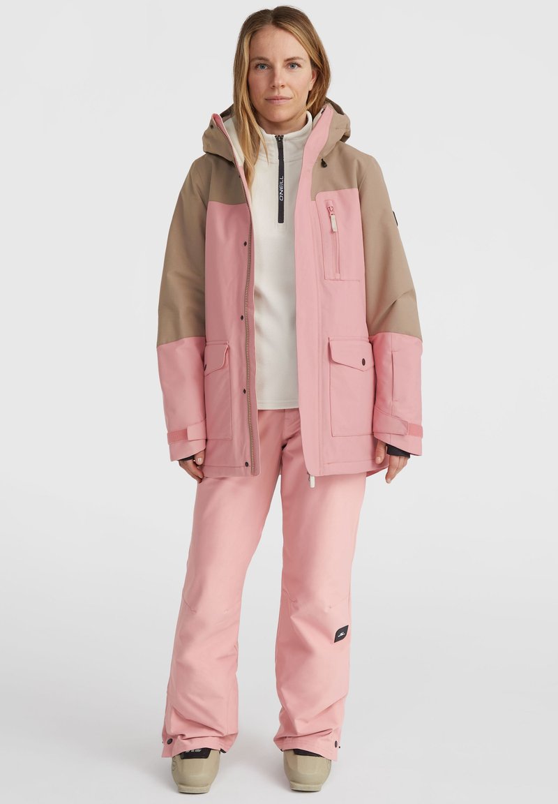 Roze en beige geïsoleerde jas met een ritssluiting aan de voorkant, grote zakken en een capuchon, gecombineerd met bijpassende roze broeken en beige laarzen. Gladde stoftextuur.