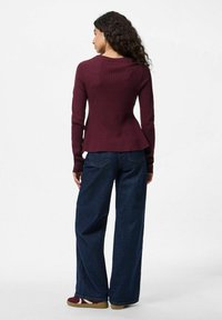 Sieviete ar lokaniem matiem, tērpusies pieguļošā bordo džemperī, platā kāju tumši zilajā biksēs un bordo sporta apavos, stāv pretējā virzienā uz vienkārša fona.