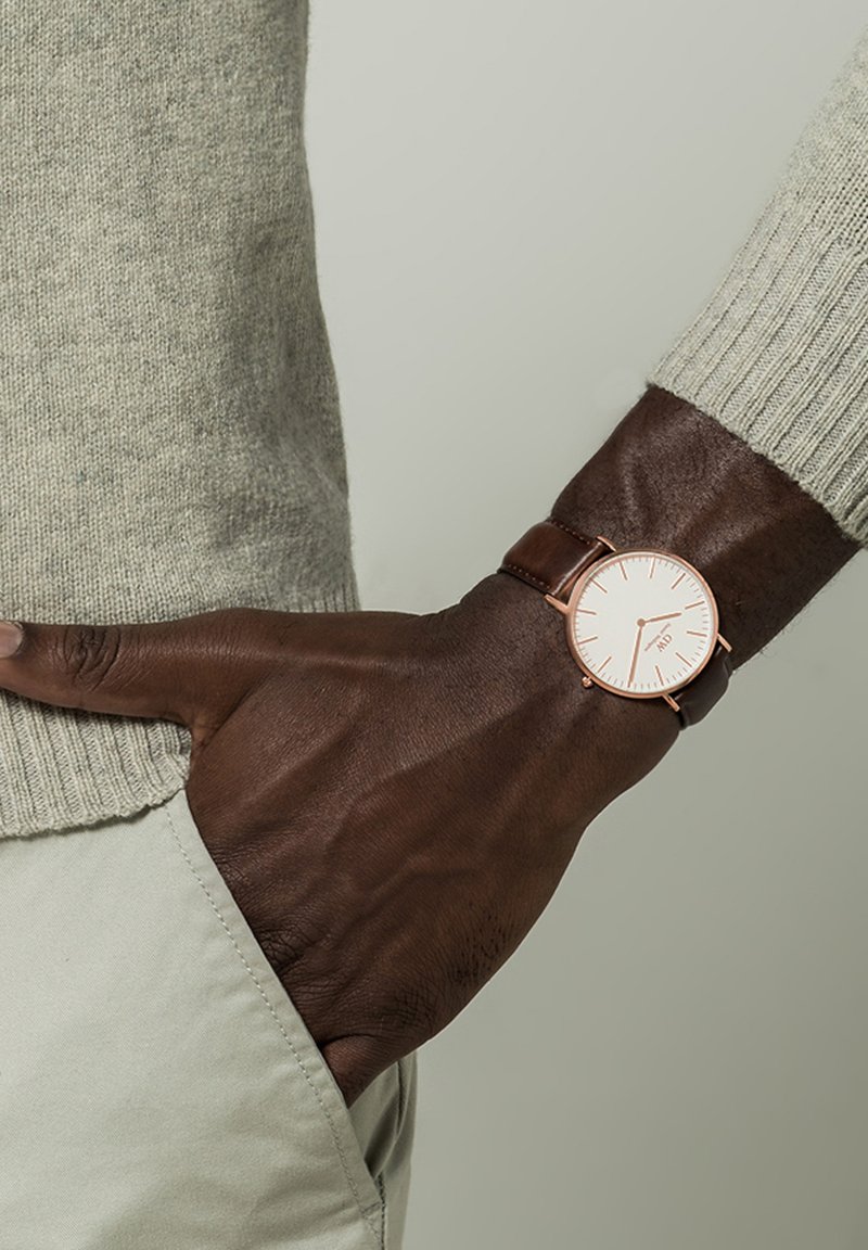 Brun läderrem klocka med en rund vit urtavla, roséguld accenter och minimala timmarkörer, som bärs på ett mörkhyat handled.