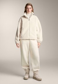 Könnyű bézs színű, texturált polár anyagból készült dzseki és nadrág szett. A dzsekin magas gallér és elülső cipzár található; a nadrágok rugalmas szegéllyel rendelkeznek.