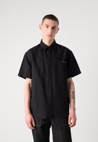 Camisa negra de manga corta con botones, hecha de una tela texturizada, que presenta un bolsillo en el pecho y un discreto detalle de logo, combinada con unos pantalones vaqueros oscuros.
