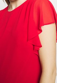 Femme portant un chemisier rouge vif avec des manches à volants superposées, montrant une partie du cou et du bras sur fond blanc.