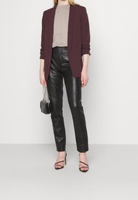 Nainen, jolla on päällään burgundinpunainen bleiseri, beige paita, mustat nahkahousut ja mustat korkokengät, joissa on hihnat. Hän pitää kädessään pientä mustaa laukkua, jossa on hopeinen ketjuhihna.