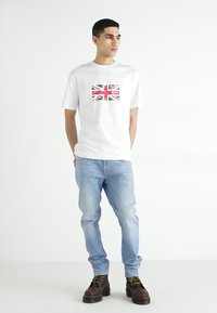Camiseta blanca de manga corta con un gráfico de la Union Jack en el pecho, combinada con jeans azul claro y botas marrones con cordones.