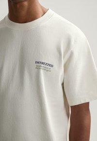 Camiseta de algodón de manga corta color crema con cuello redondo acanalado y texto bordado en azul marino: "DSTREZZED Hermandad del gont moderno."