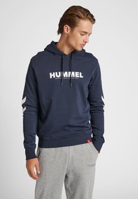 Man i marinblå hoodie med vit "HUMMEL"-logga och grå mjukisbyxor, stående med händerna i fickorna och tittar ner åt vänster.
