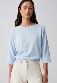 Lichtblauwe, gestructureerde top met een brede halslijn en 3/4 mouwen, gecombineerd met lichtgekleurde broek met een knoop en zijzakken.