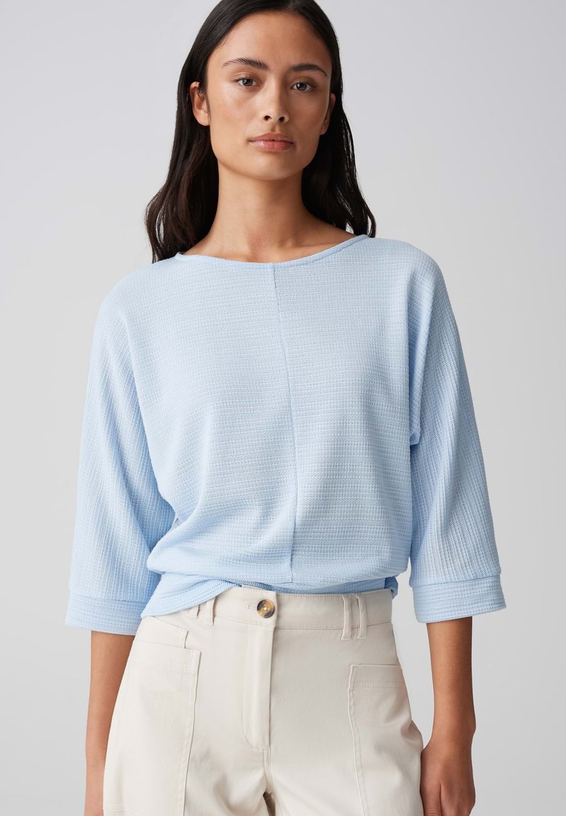 Lichtblauwe, gestructureerde top met een brede halslijn en 3/4 mouwen, gecombineerd met lichtgekleurde broek met een knoop en zijzakken.
