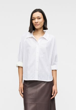 Cămașă albă cu nasturi, cu guler și mâneci suflecate, având un design texturat. Asortată cu o fustă din piele maro închis.