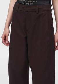 Personne portant un pantalon large marron foncé et un haut noir avec un texte blanc à la taille, mains détendues sur les côtés.