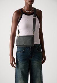 Borsa a tracolla nera in pelle con finitura texture, forma rettangolare, accessori in oro e tracolla regolabile, indossata sopra un top leggero e dei jeans.