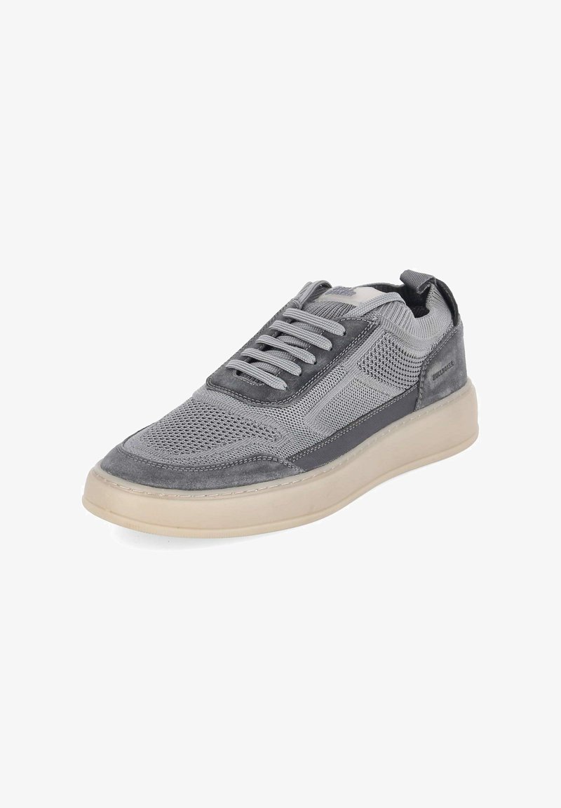 Zapatilla gris con parte superior de malla y ante, cierre con cordones y suela gruesa de color blanco roto, inclinada para mostrar detalles laterales y superiores.