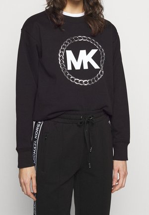 Person trägt ein schwarzes Langarmshirt mit großem weißem „MK“-Logo in einem silbernen Kettenkreis, kombiniert mit schwarzen Hosen mit Kordelzug und weißem Seitentext.