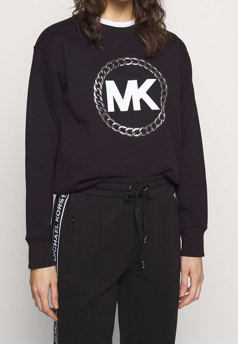 Personne portant un t-shirt noir à manches longues avec un grand logo blanc "MK" à l'intérieur d'un cercle en chaîne argentée, associé à un pantalon noir à cordon avec un texte blanc sur le côté.