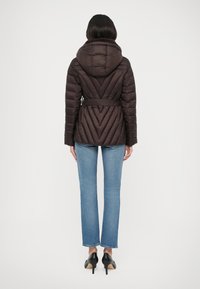 Doudoune marron avec une taille ceinturée, un motif en losanges et une capuche. Portée sur un jean évasé bleu et des talons noirs.