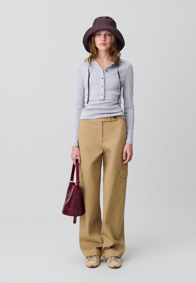 Maglione grigio a maniche lunghe con bottoni, pantaloni beige a gamba larga con tasche, cappello a secchiello marrone e borsa a spalla bordeaux. Sneakers marroni e beige.