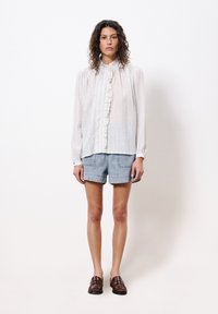 Weiße Bluse mit Rüschen vorne, gestreiftes Design; kombiniert mit hellblauen Jeansshorts. Das Model trägt braune Lederschuhe.