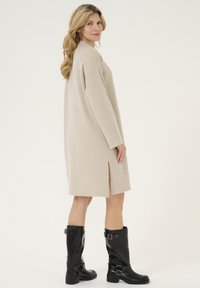 Robe beige côtelée à manches longues, longueur genou et fentes sur les côtés. Assorti à des bottes noires montant jusqu'aux genoux avec des boucles et une finition lisse.