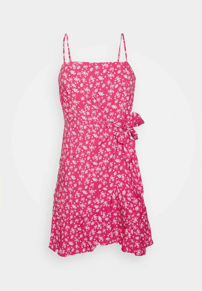 Robe rose sans manches avec imprimé floral blanc, fines bretelles, taille cintrée nouée sur le côté en nœud, et un ourlet à volants.