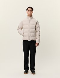 Casaco puffer bege com colarinho alto e fecho de correr frontal, usado sobre calças pretas. O casaco apresenta um design acolchoado e um corte descontraído.
