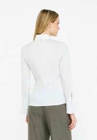 Weiße Bluse mit langen Ärmeln, hohem Kragen, gepufften Manschetten und taillierter Silhouette. Verfügt über einen dezenten Rückenverschluss und eine glatte Textur.