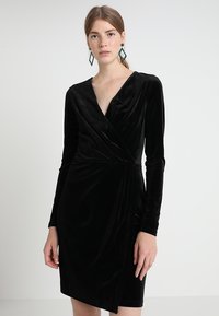 Rochie din catifea neagră, cu mâneci lungi, decolteu adânc în formă de V și o siluetă ajustată. Dispune de o fanta laterală pentru un aspect modern.