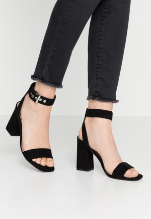 Fødder iført sorte ruskindssandaler med blokhæl, ankelrem og sølvspænde, kombineret med sorte skinny jeans med frynset kant.