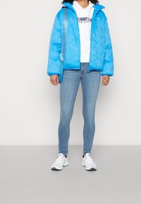 Chaqueta puffer azul con estampado de logotipo, capucha y puños elásticos, llevada sobre una sudadera blanca, combinada con jeans ajustados de un azul claro y zapatillas.