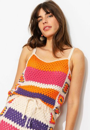 Femme aux cheveux bruns portant un haut crochet coloré et une jupe assortie avec des motifs orange, rose, violet et blanc, posant devant un fond blanc.