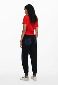 Rode t-shirt met korte mouwen, gecombineerd met zwarte joggers met denim accenten en zakken; elastische mouwen; model staat met de rug naar de camera en toont de details aan de achterkant.