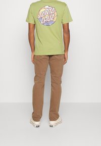 Zelené bavlněné tričko s kruhovou vícebarevnou grafikou na zádech, doplněné hnědými kalhotami a bílými teniskami, pohled zezadu.