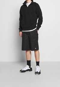 Persoon staande in een zwart kwart-ritssweater, zwarte knielange shorts, zwarte sokken en zwart-witte sneakers tegen een effen achtergrond.